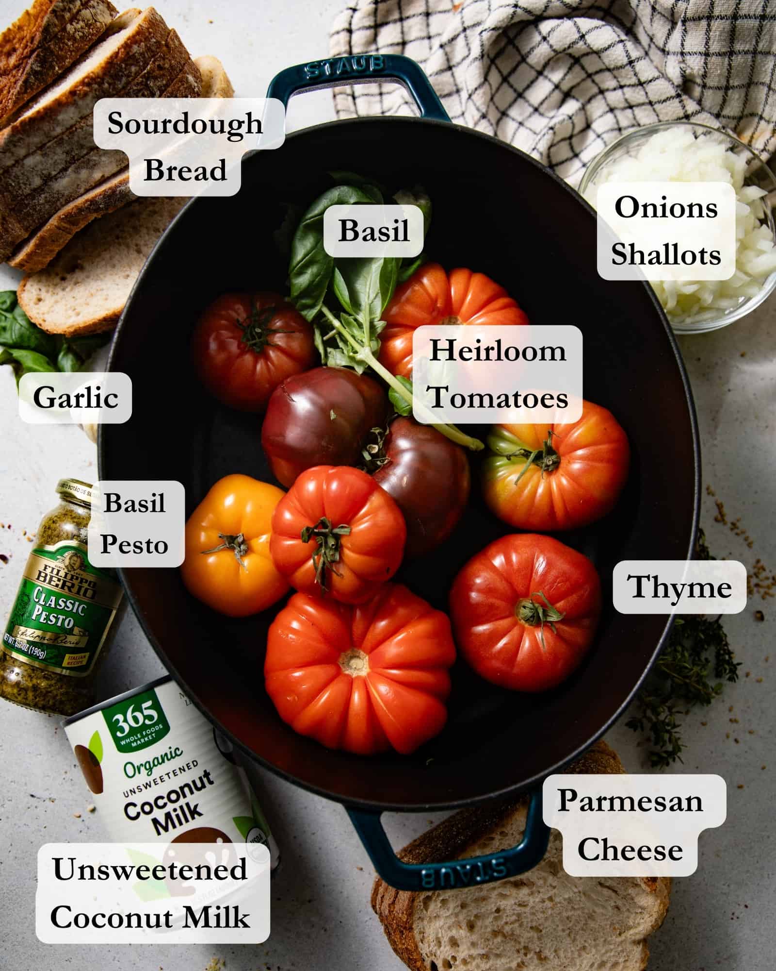 ingredients to make tomato soup in bowl on a white table - heirloom tomatoes, thyme, basil, garlic, pesto, coconut milk, bread, olive oil, onions, and shallots