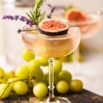a lavender cocktail in a coupe glass garnished with rosemary, fresh lavender, and a fig.