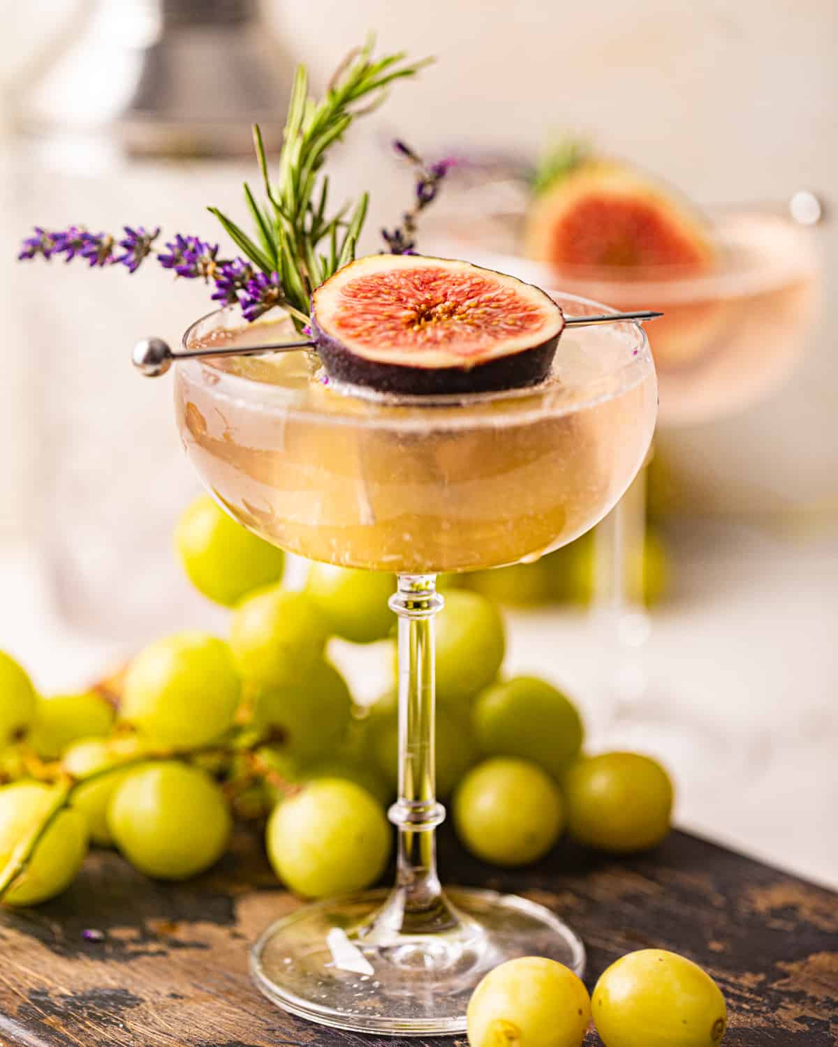 a lavender cocktail in a coupe glass garnished with rosemary, fresh lavender, and a fig.