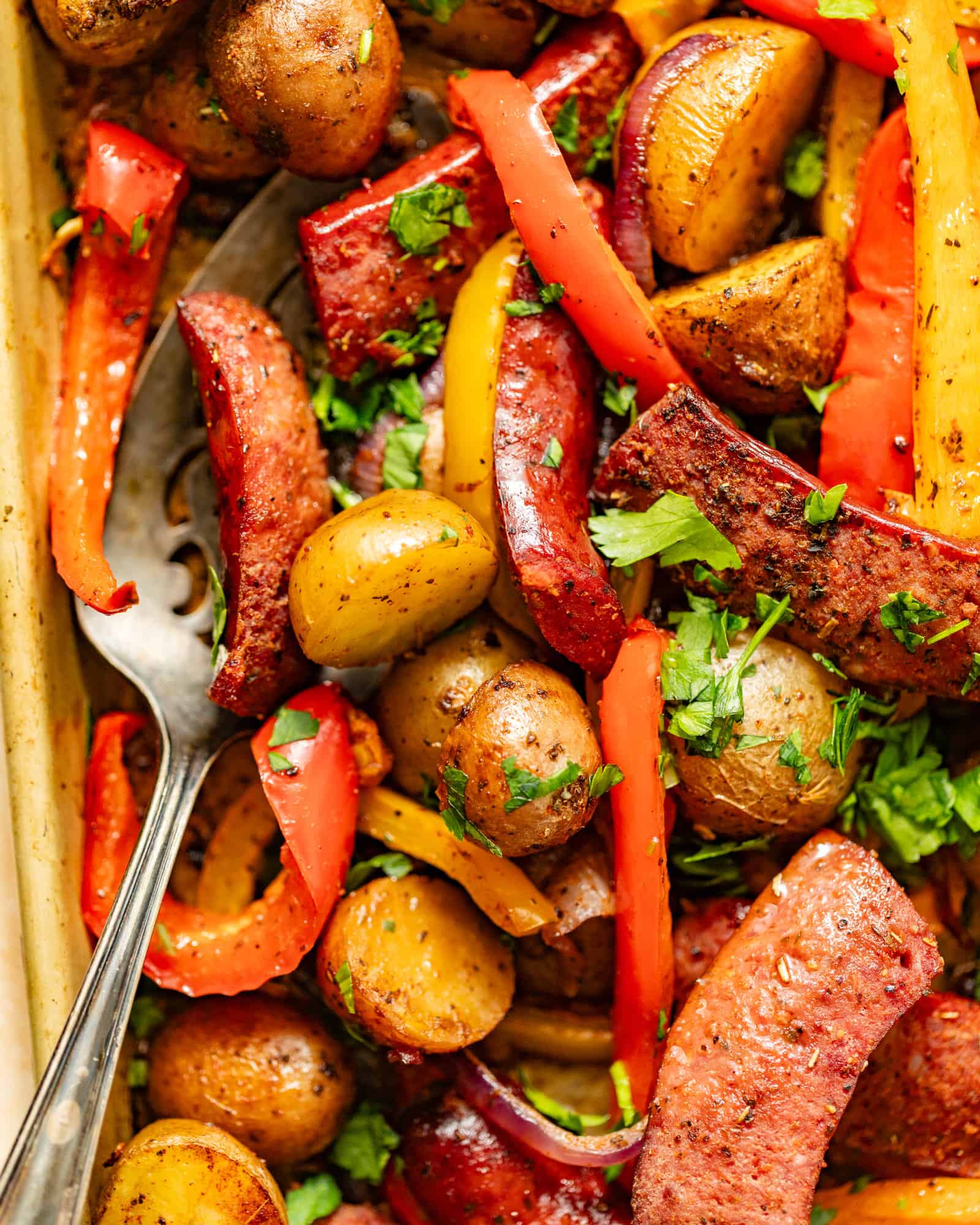 sausages peppers onions and potatoes on a sheet pan with fresh parsley on top.