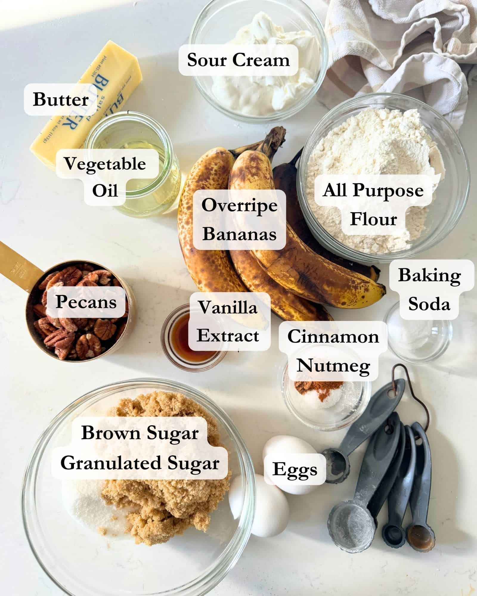 ingredients to make banana bread on a white surface - overripe bananas, sugar, brown sugar, eggs, flour, cinnamon, nutmeg, sour cream, vanilla extract, pecans, butter, vegetable oil, and baking soda