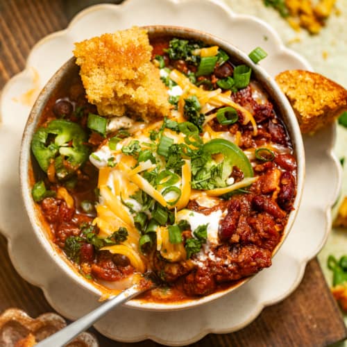 a bowl of chili on a plate topped with jalapenos, sour cream, tortilla chips, and cheese.