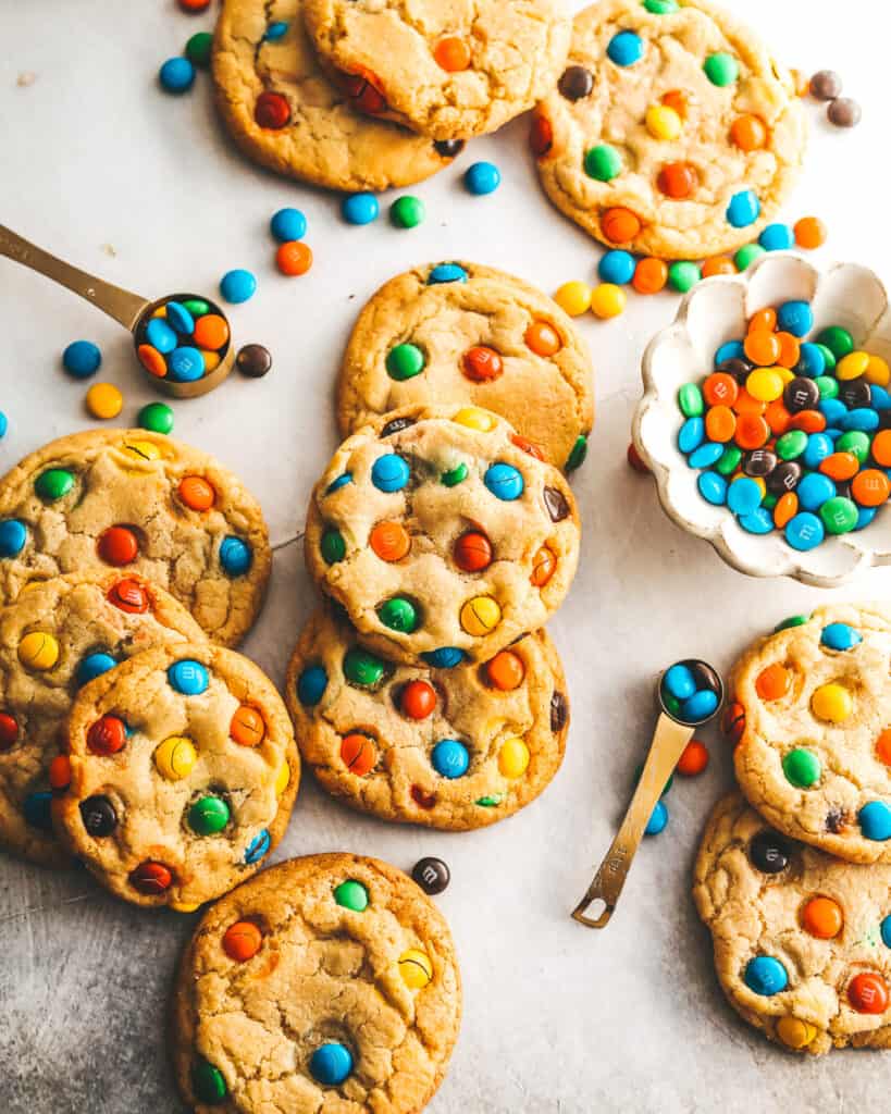 Chewy M&M Cookies laid on a flat surface with M&Ms in a bowl