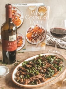 A plate of skirt steak with a cookbook in the background and a wine bottle and wine in wine glasses