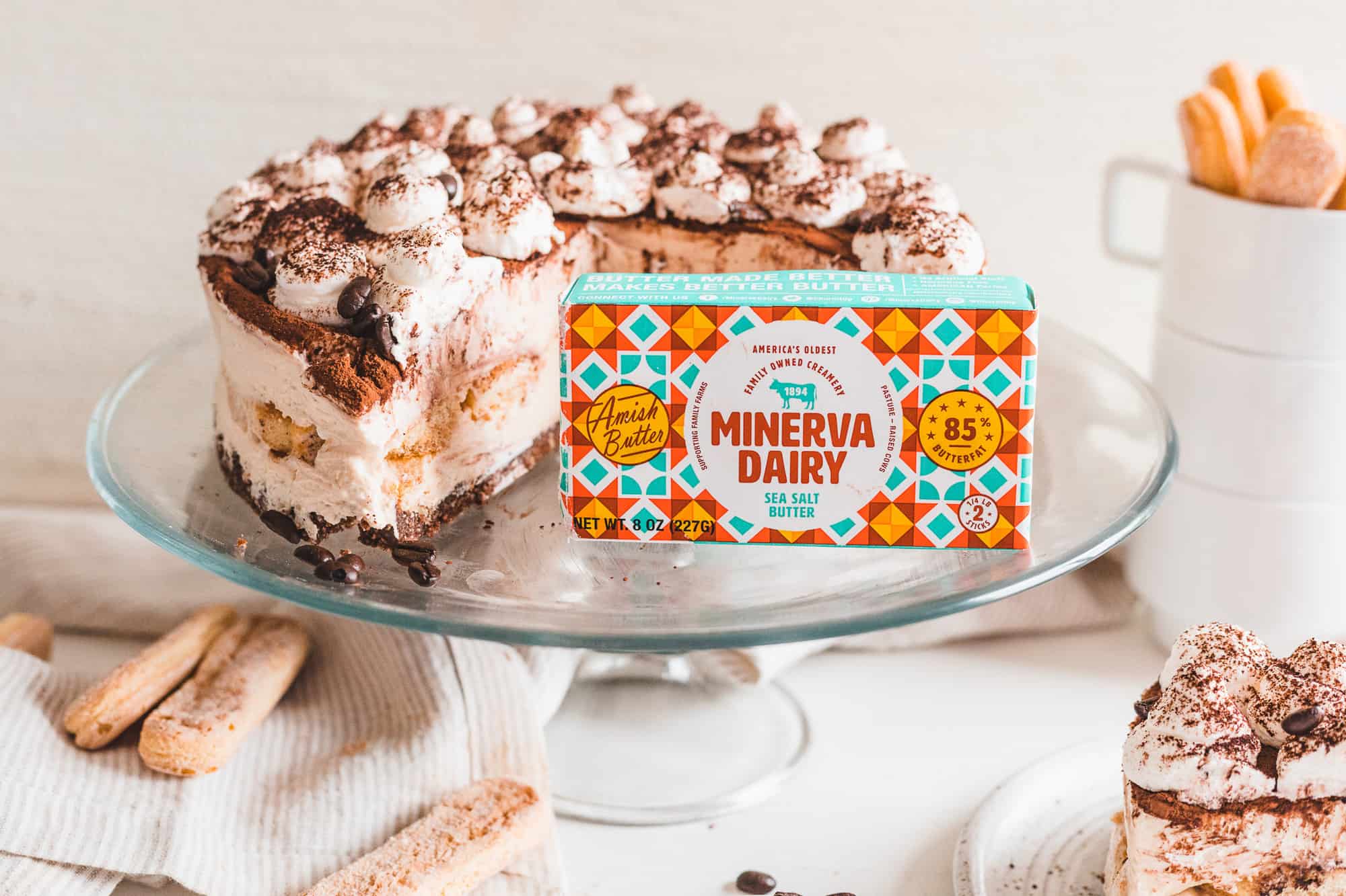 A sliced No-Bake Tiramisu Cheesecake on an elevated serving dish with an 8 ounce pack of Minerva Dairy Sea Salt Butter at the forefront.