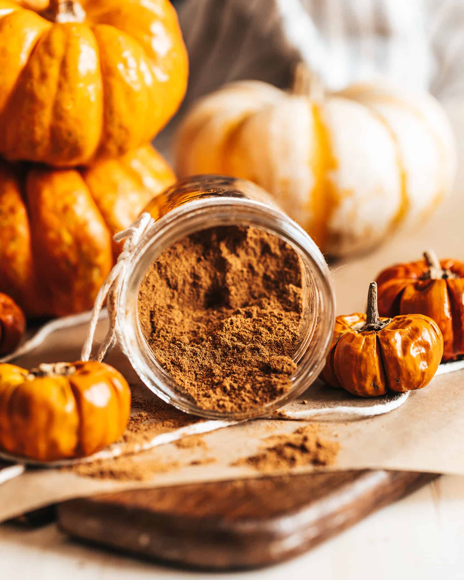 An open jar of Homemade Pumpkin Spice with some seasoning spilled on the table with an assortment of pumpkins in the background.