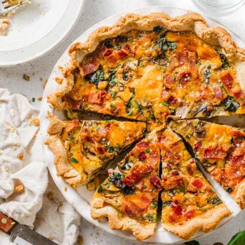 bacon mushroom spinach quiche in a pie dish with fresh herbs and lemons in a bowl next to it.