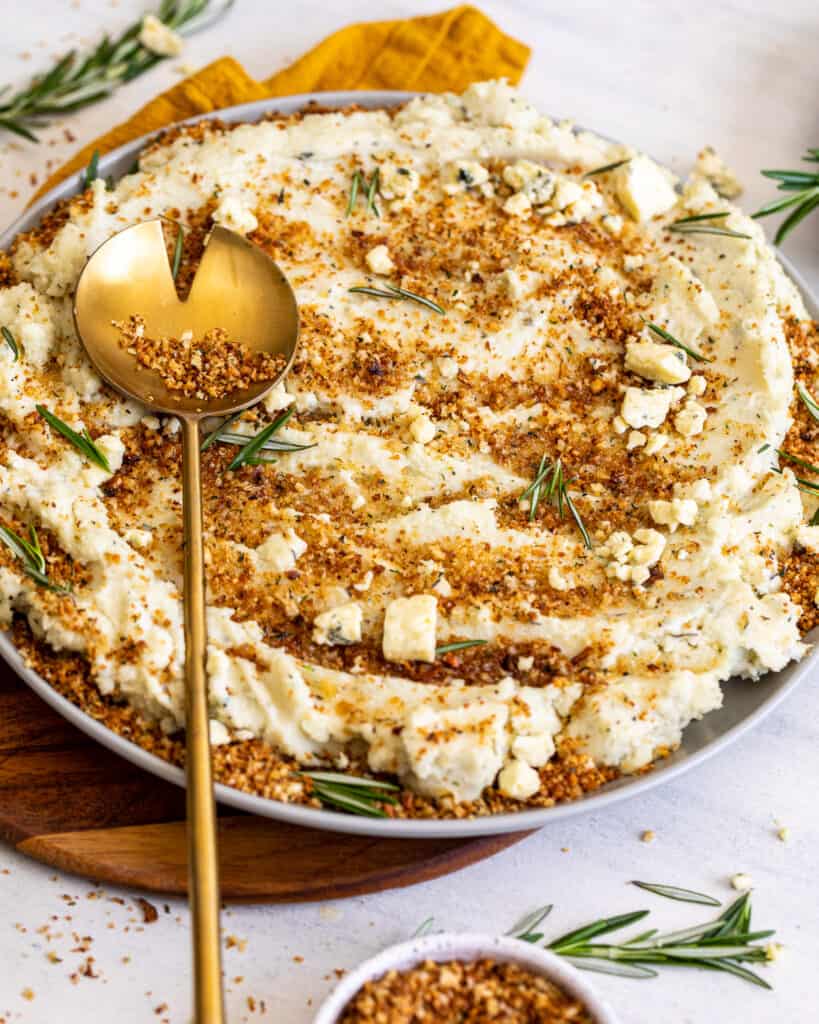 A gold spoon placed over a large bowl of Blue Cheese Mashed Potatoes with Toasted Breadcrumbs.