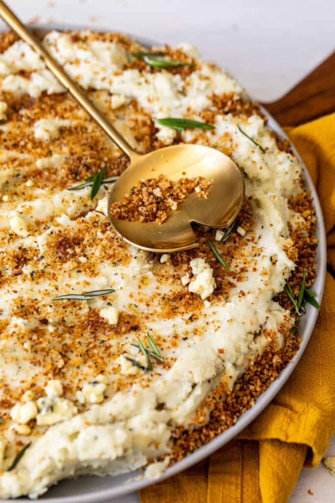 A gold spoon placed over a large bowl of Blue Cheese Mashed Potatoes with Toasted Breadcrumbs.