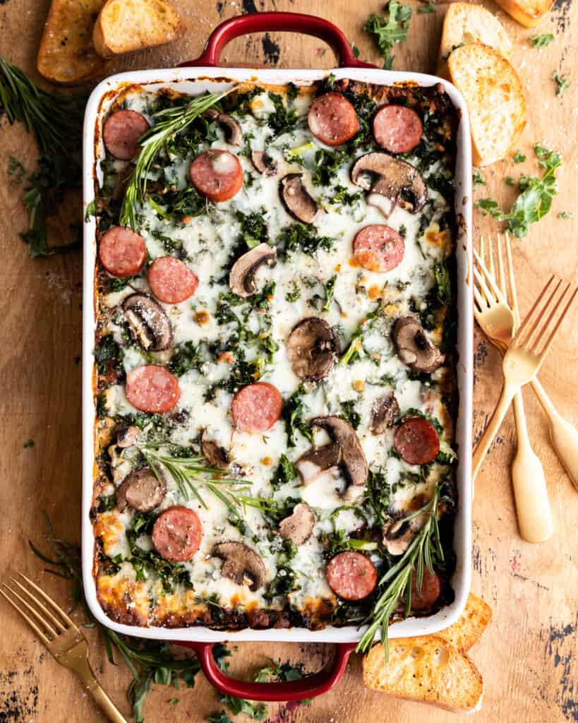 sausage kale and mushroom lasagna in a baking dish next to forks and small pieces of bread