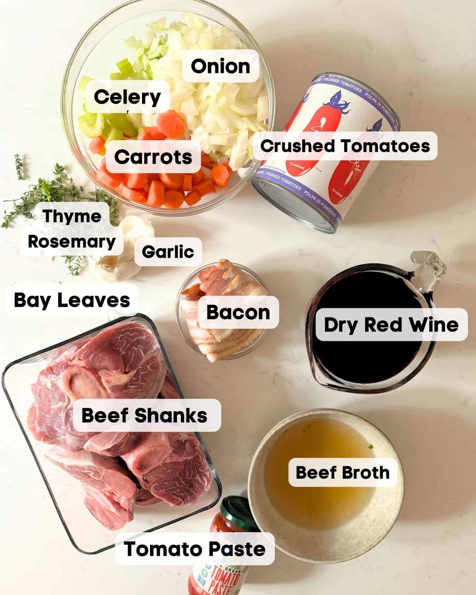 ingredients to make osso buco on a white surface - beef shanks, red wine, garlic, hersb, bacon, beef broth, tomato paste, crushed tomatoes, celery, carrots, and onions.