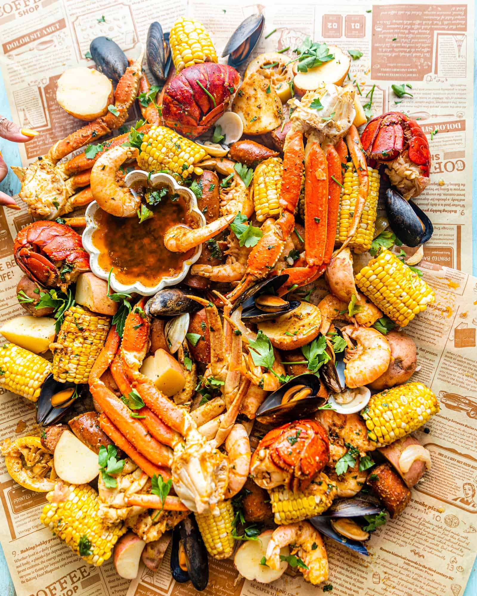 Shrimp, Lobster, Crab Legs, Clams, Corn, Mussels, and Sausages on a piece of newspaper with garlic butter sauce