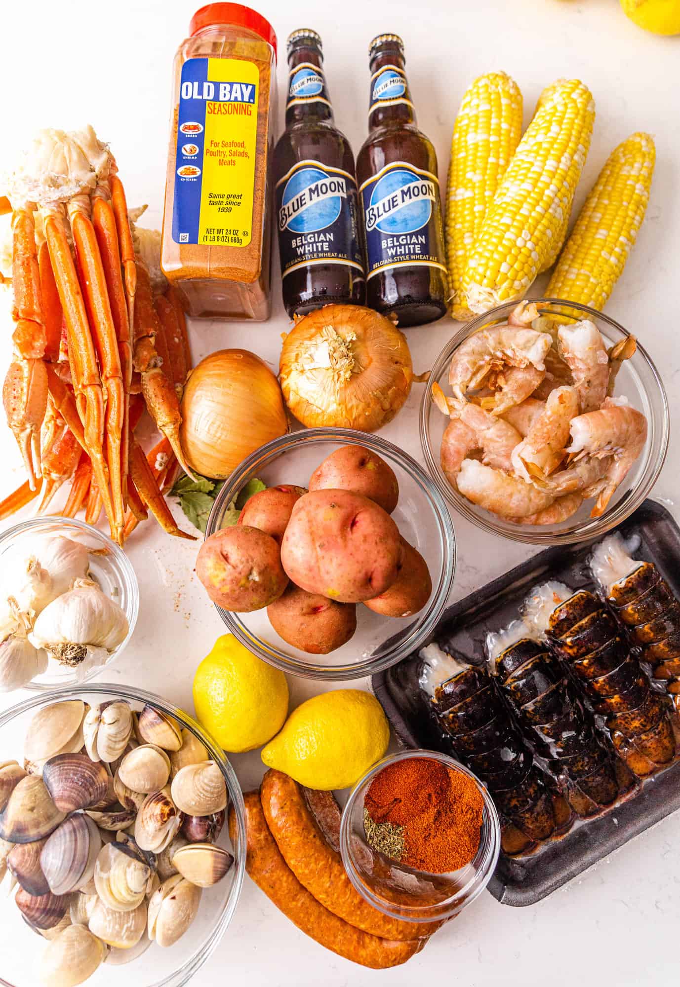 ingredients to make a cajun seafood boil on a white surface - beer, old bay seasoning, corn, shrimp, onions, lobster, smoked sausage, crab legs, potatoes, mussels, lemons, garlic, bay leaves, cajun seasoning, and clams.