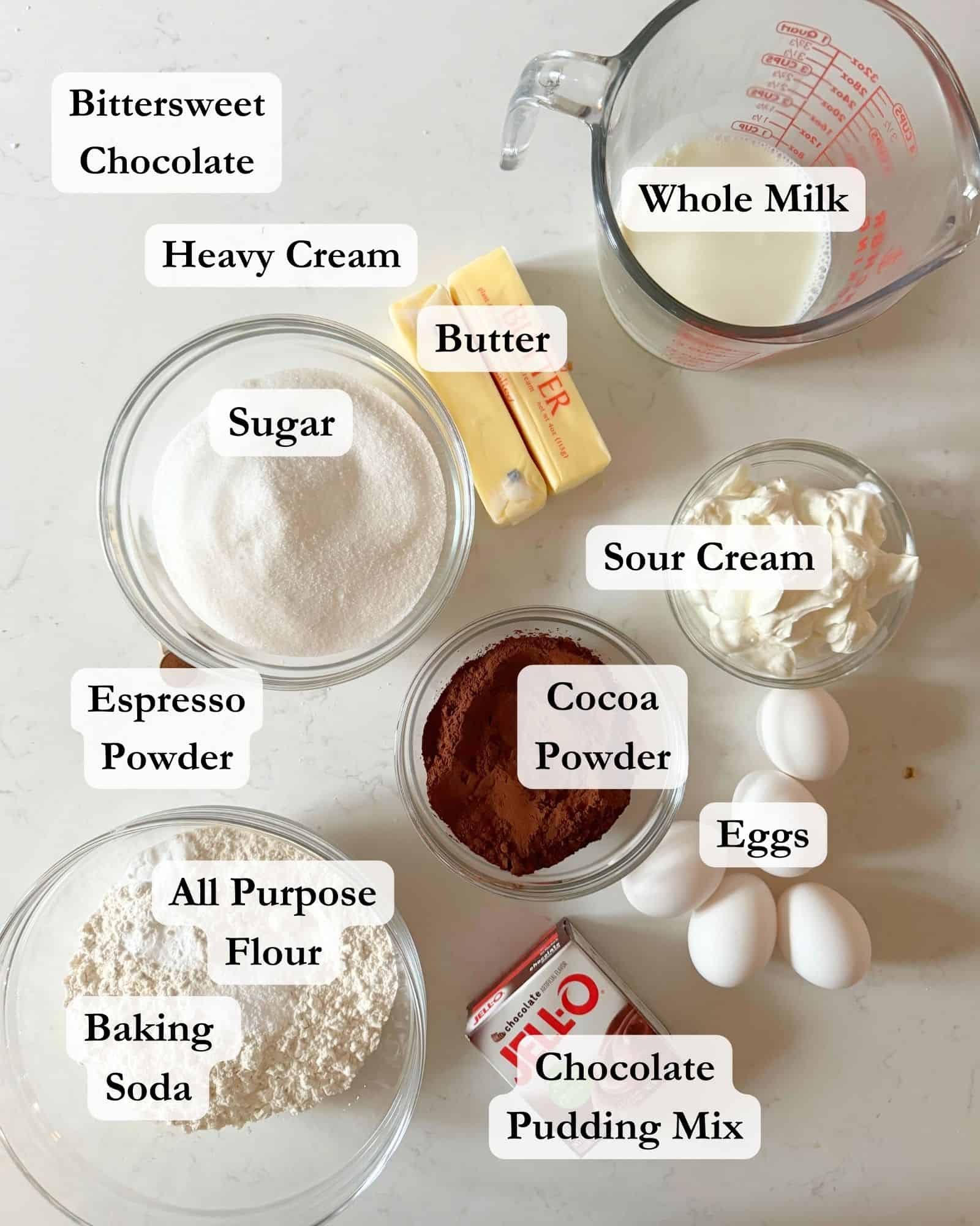 chocolate pound cake ingredients on a white surface in small bowls. Flour, sugar, butter, pudding mix, eggs, baking soda, espresso powder, cocoa powder, bittersweet. chocolate, heavy cream, sour cream, and whole milk