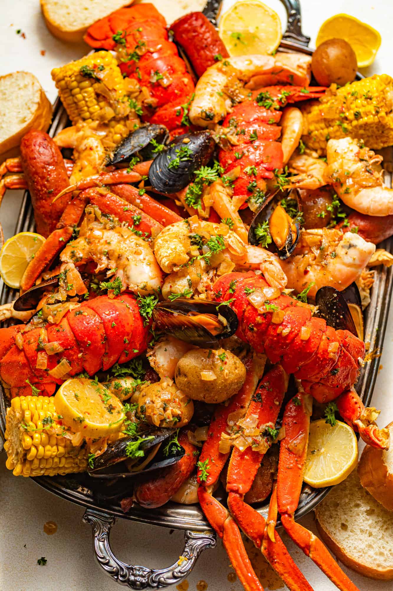 crabs legs, lobster, corn, sausage, potatoes, clams, mussels, and shrimp all on a silver serving tray covered in seafood boil sauce and fresh parsley