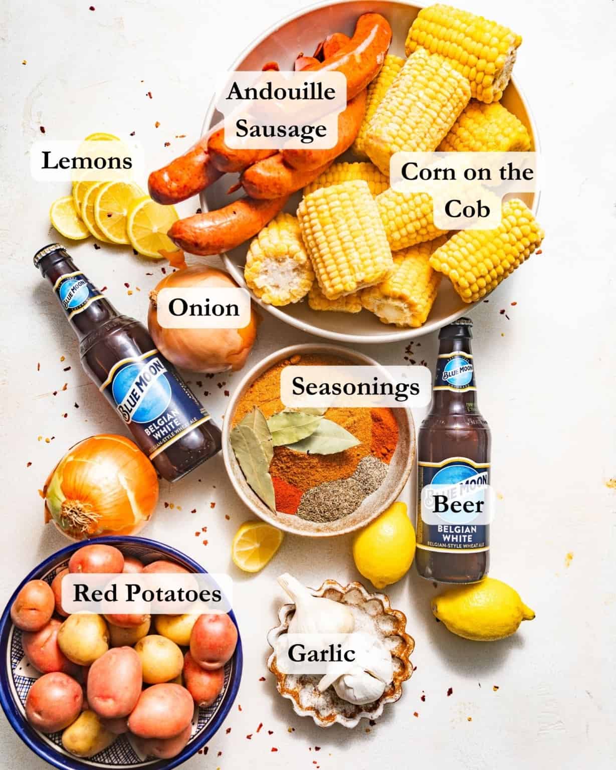 ingredients to make a cajun seafood boil on a white surface - beer, old bay seasoning, corn, onions, smoked sausage, potatoes, lemons, garlic, bay leaves, and cajun seasoning.