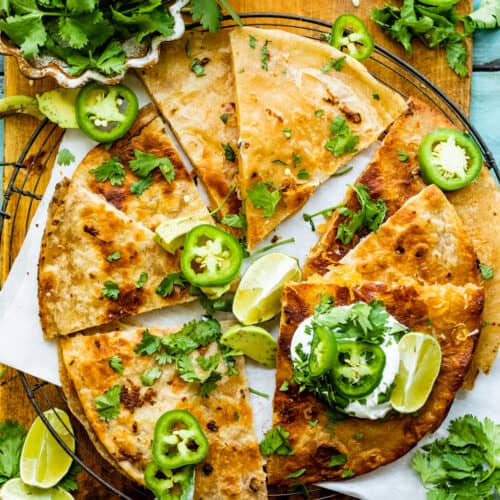 cheese quesadilla on a serving board garnished with limes, jalapenos, and cilantro.
