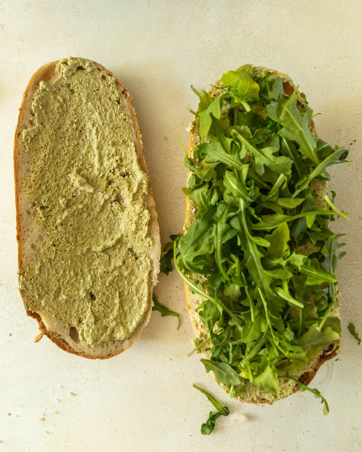 cibatta bread sliced in half. One side has pesto mayo spread on top and the other side of bread has arugula on top of the bread.