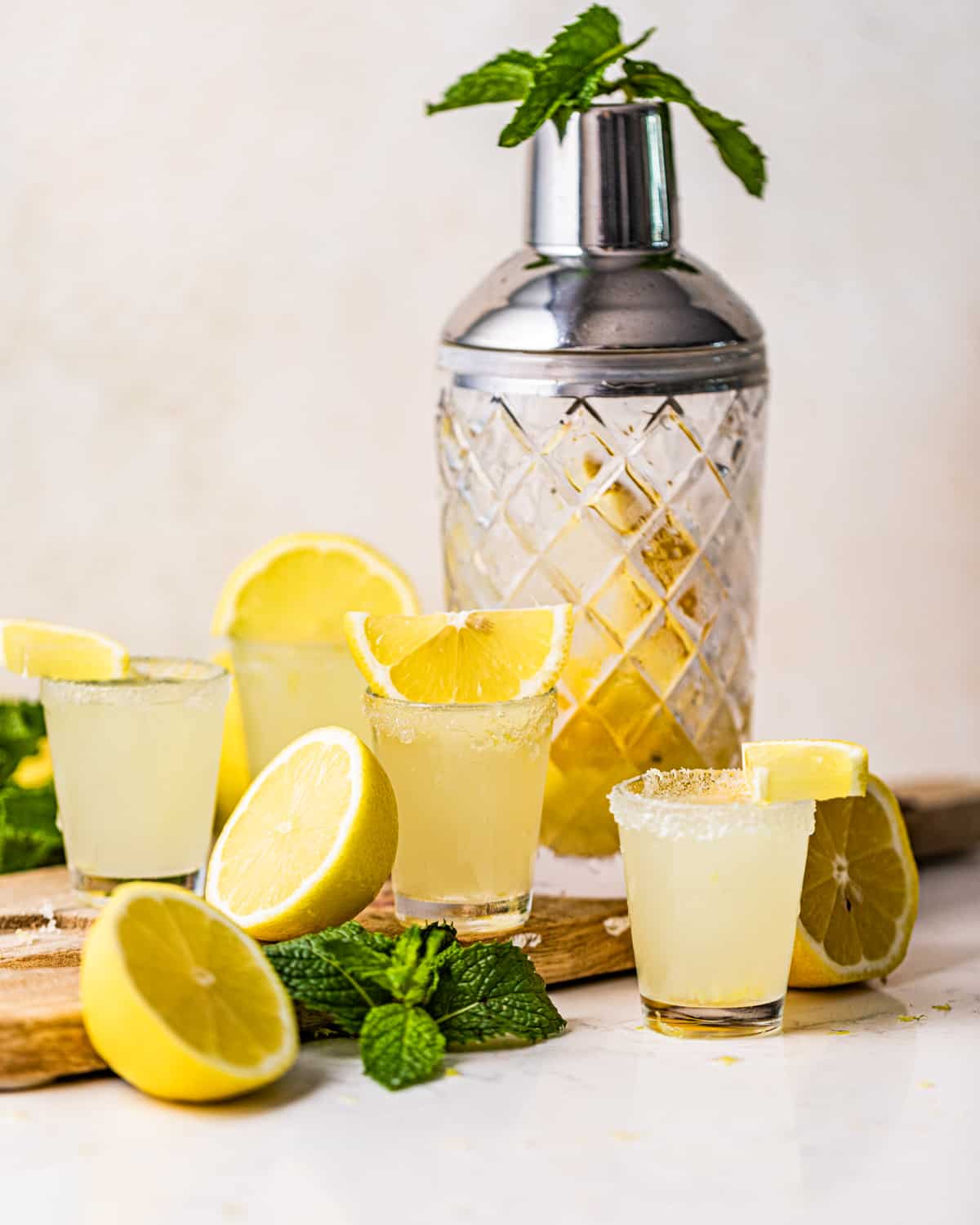 lemon drop shots on a serving board with lemon wedge, mint leaves, and a cocktail shaker.