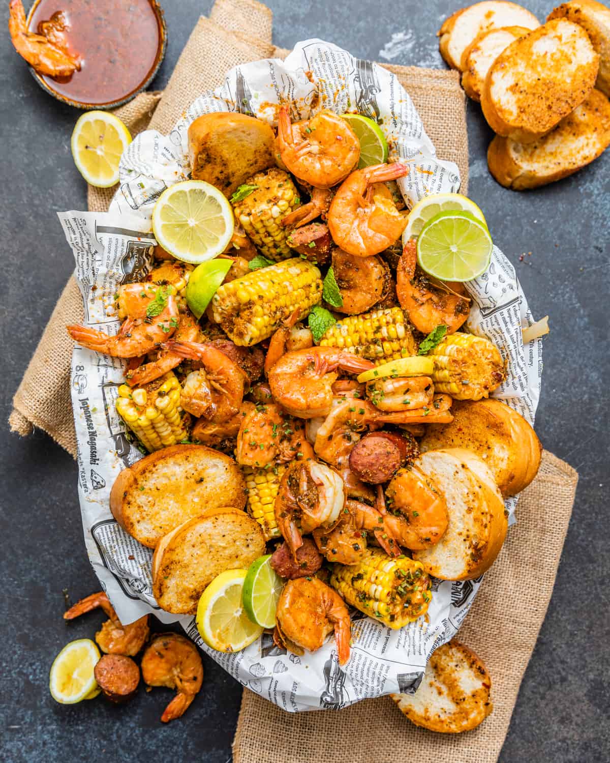 low country boil in a oval platter on top of newspaper surrounded by shrimp and sliced bread.