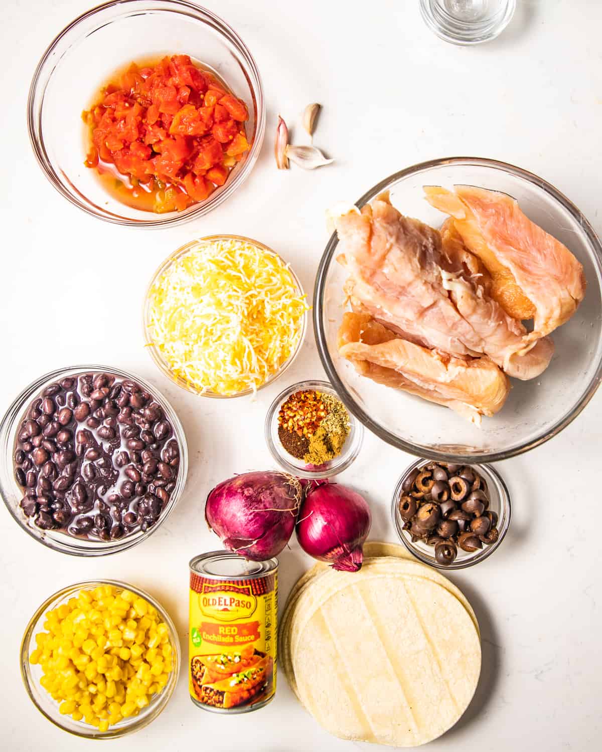 Ingredients to make chicken enchiladas - tomatoes, chicken, red onion, black beans, corn, seasonings, tortillas, enchilada sauce, olives, and cheese.