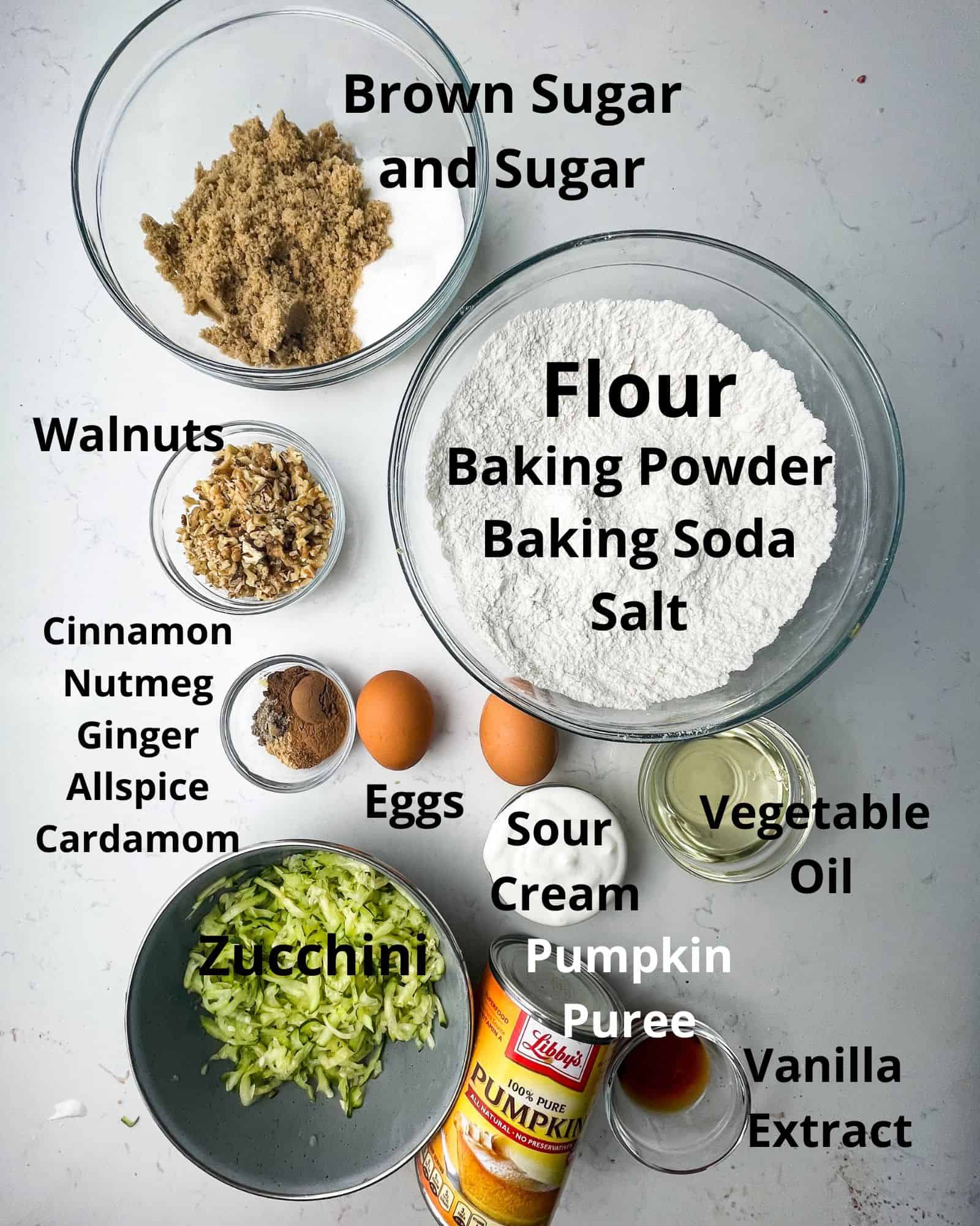 ingredients to make zucchini pumpkin muffins - flour, baking soda, baking powder, salt, cinnamon, nutmeg, ground ginger, ground nutmeg, allspice, cardamom, brown sugar, sugar, vegetable oil, sour cream, eggs, vanilla extract, pumpkin puree, zucchini, and walnuts.