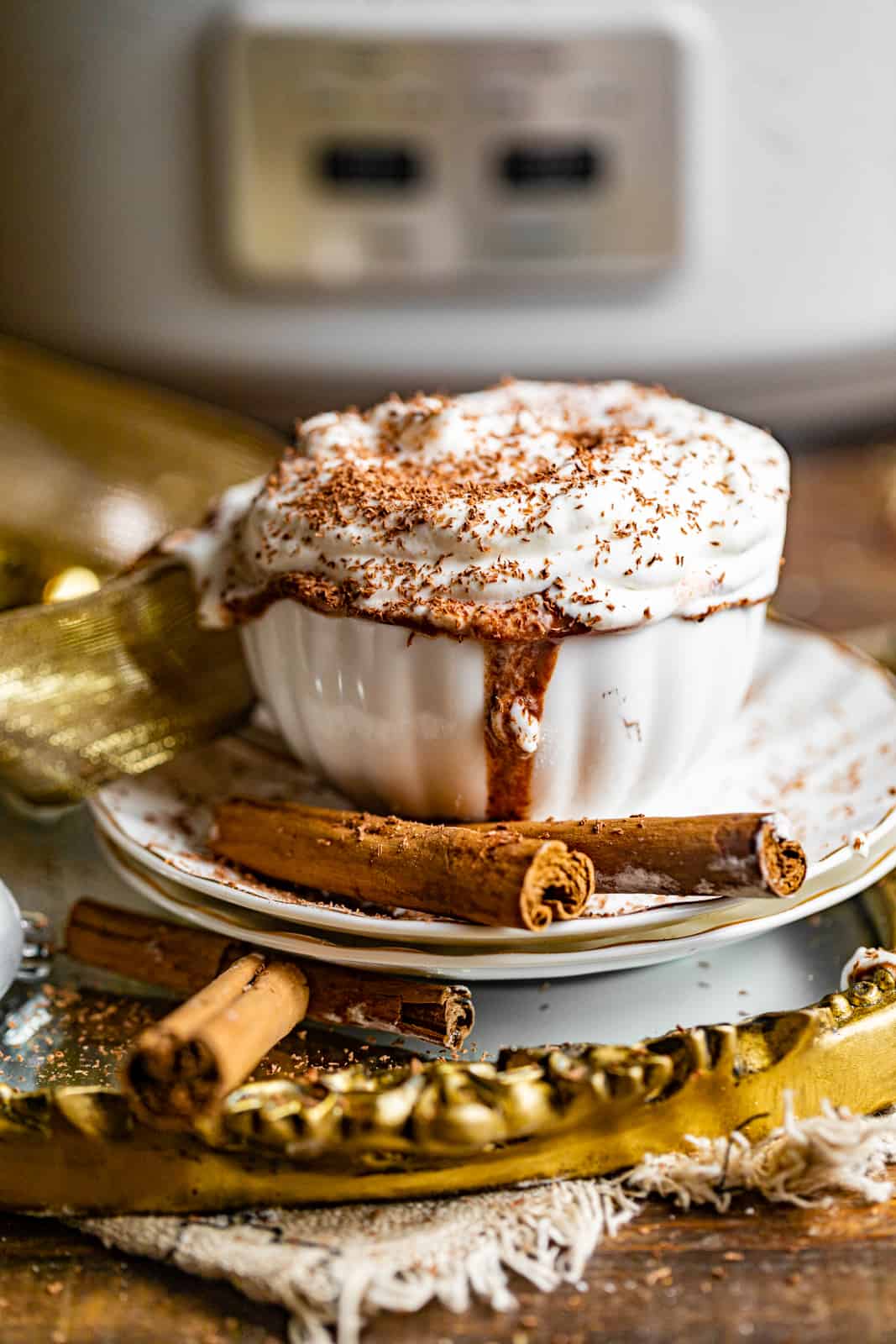 a cup of hot chocolate in front of a crockpot slow cooker garnished with whipped cream and shaved chocolate.