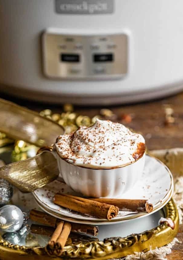 a cup of hot chocolate in front of a crockpot slow cooker garnished with whipped cream and shaved chocolate.