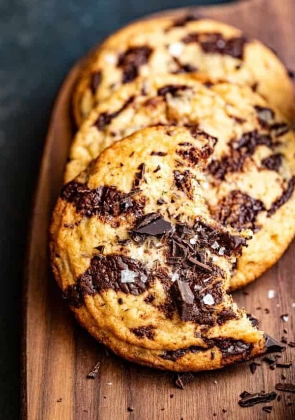 three chocolate chip cookies on a brown wooden board with chopped chocolate and flake salt on top.