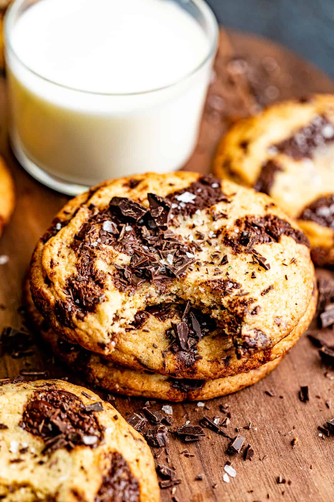 chocolate chip cookies stacked on top of each other in front of a glass of milk garnished with flake salt and chocolate chunks.