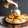 White gravy in a gravy boat being poured on top of a plate of of biscuits.