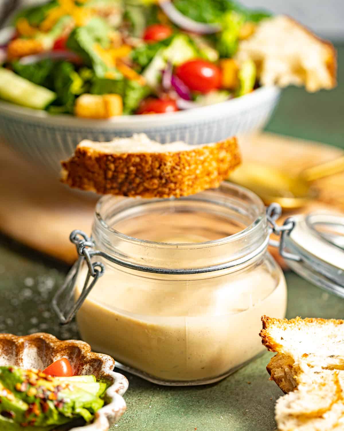 creamy balsamic dressing in a jar in front of a bowl of salad with a slice of bread on top.