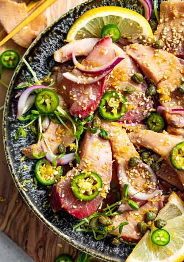 hamachi crudo on a plate with lemons, capers, and sliced peppers and red onions.