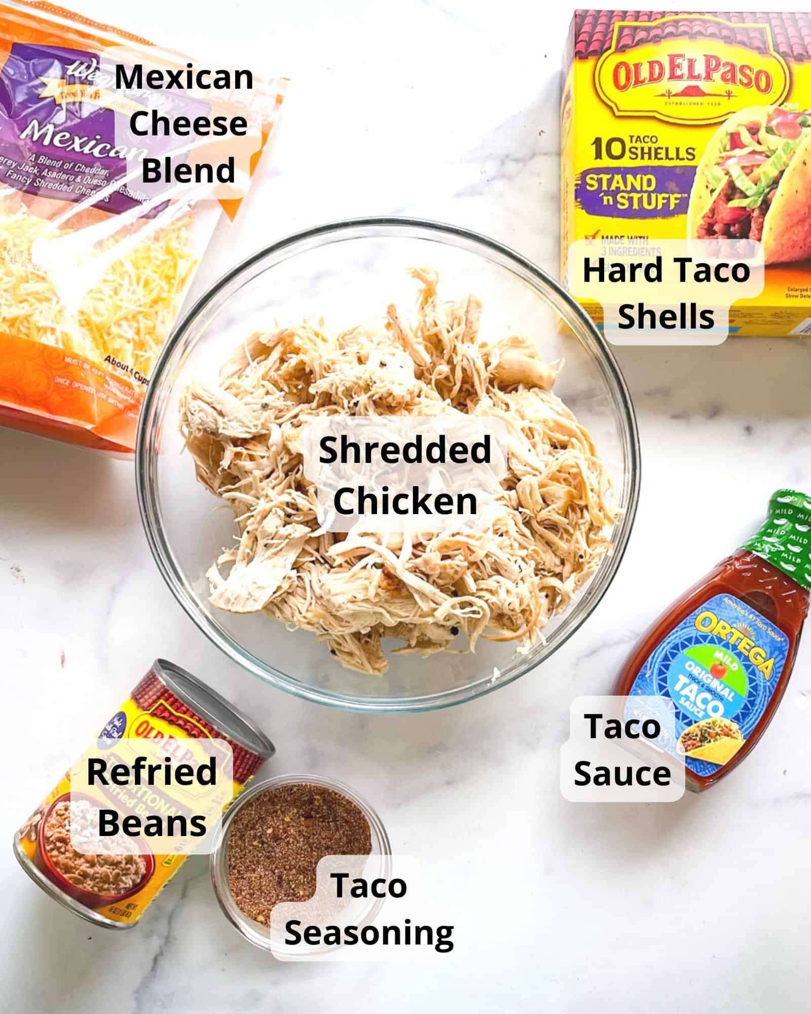 ingredients to make chicken tacos - shredded chicken, taco sauce, mexican cheese blend, refried beans, hard taco shells, and taco seasoning.