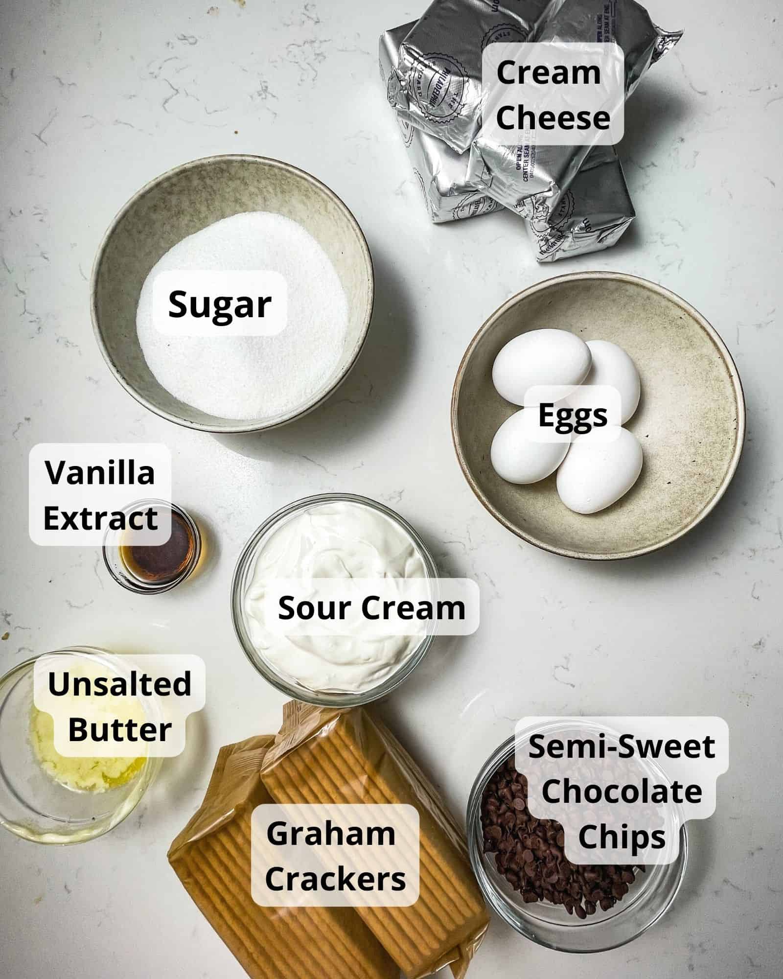 ingredients to make chocolate chip cheesecake - cream cheese, sugar, eggs, vanilla extract, sour cream, melted butter, graham crackers, chocolate chips, and heavy cream.