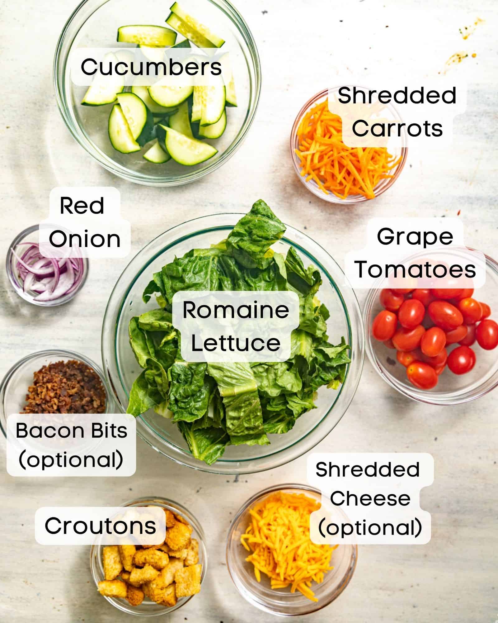 ingredients to make a house salad - lettuce, cucumbers, shredded carrots, red onion, tomatoes, bacon bits, shredded cheese, and croutons.