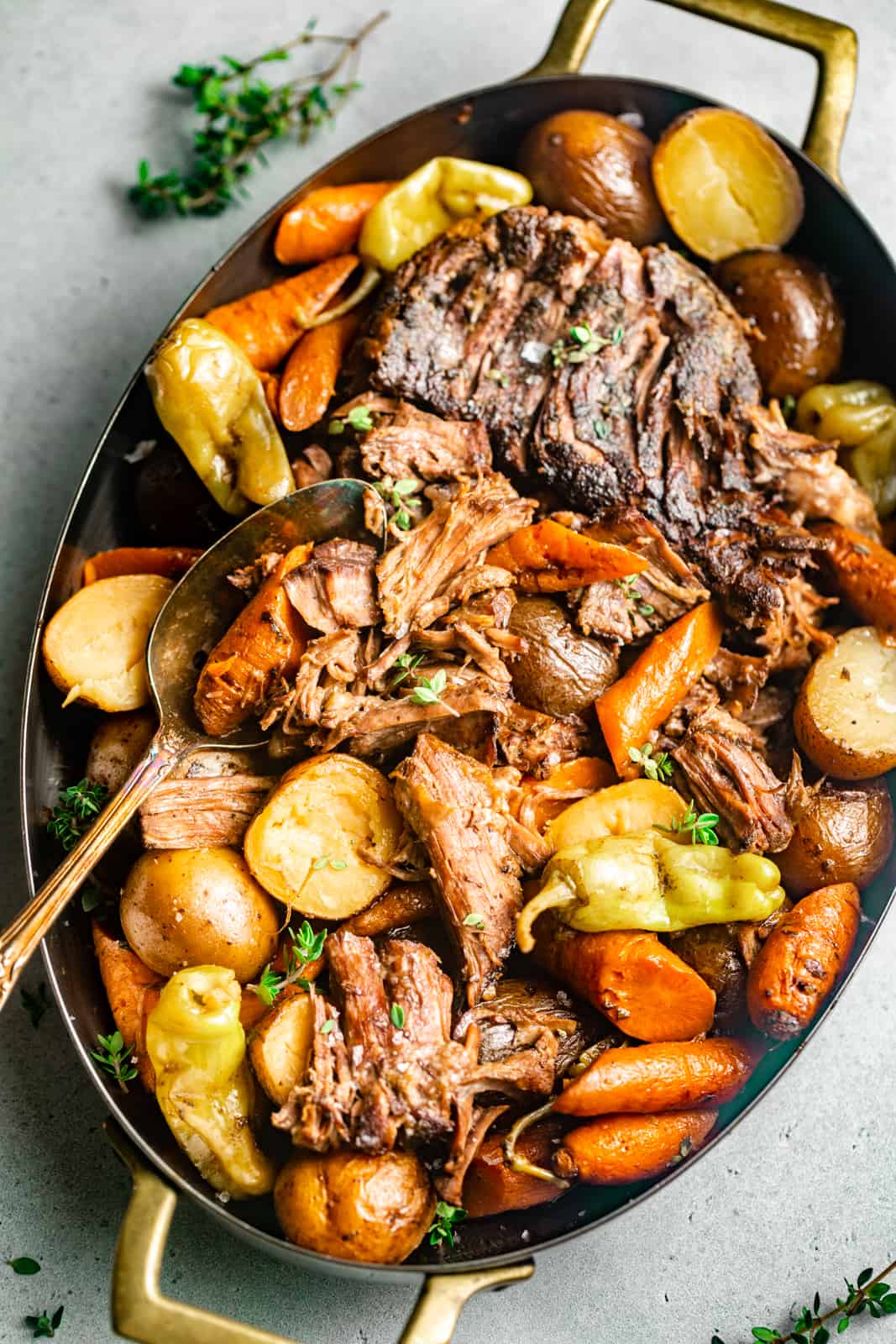 mississippi pot roast in a serving dish with a spoon and garnished with fresh thyme and pepperoncini peppers.
