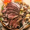 sliced smoked leg of lamb on a wooden plate with potatoes and fresh herbs.