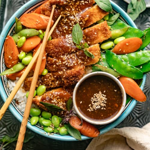teriyaki chicken, edamame beans, carrots, snow peas, and rice in a large bowl with chopsticks and teriyaki sauce.