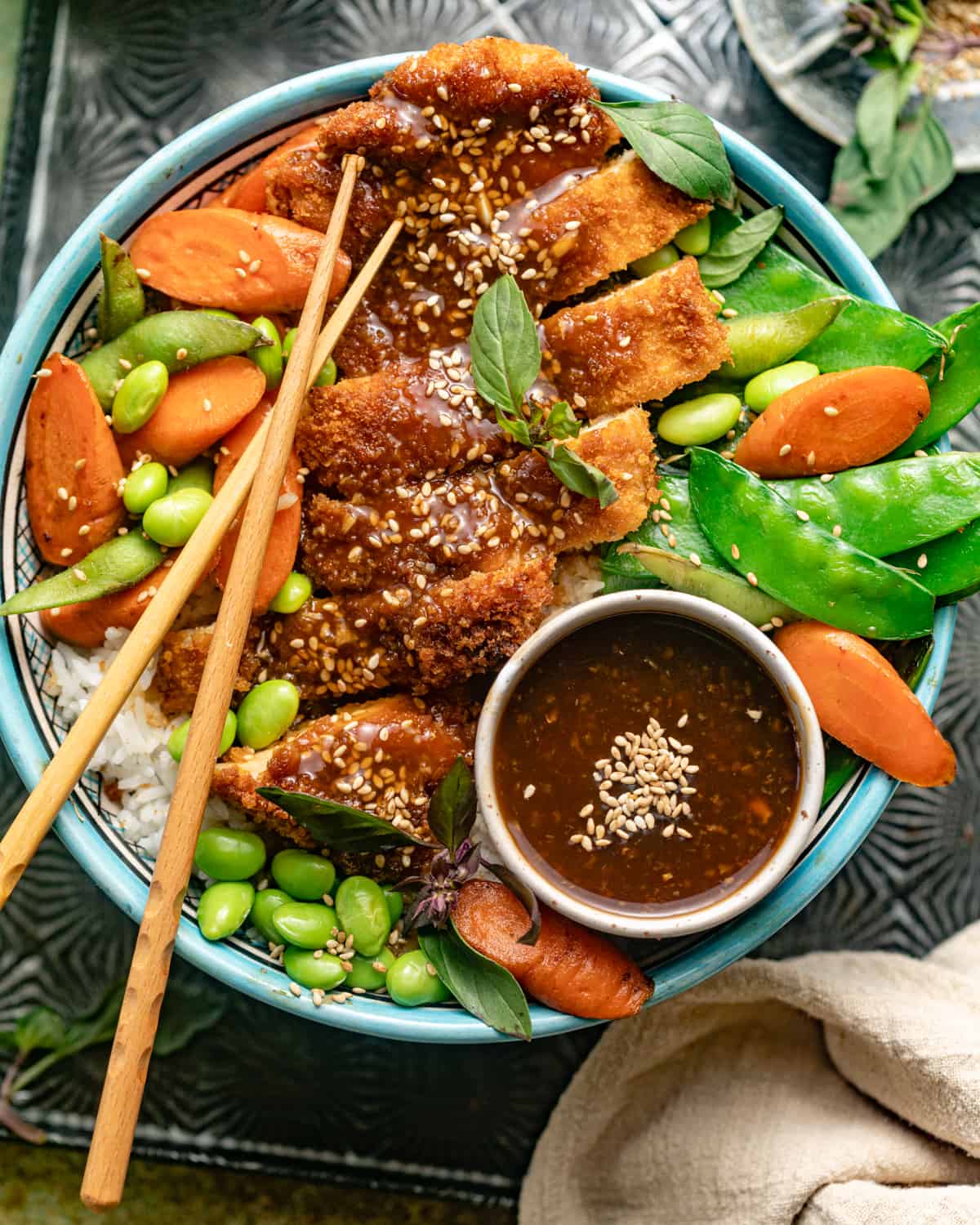 teriyaki chicken, edamame beans, carrots, snow peas, and rice in a large bowl with chopsticks and teriyaki sauce.