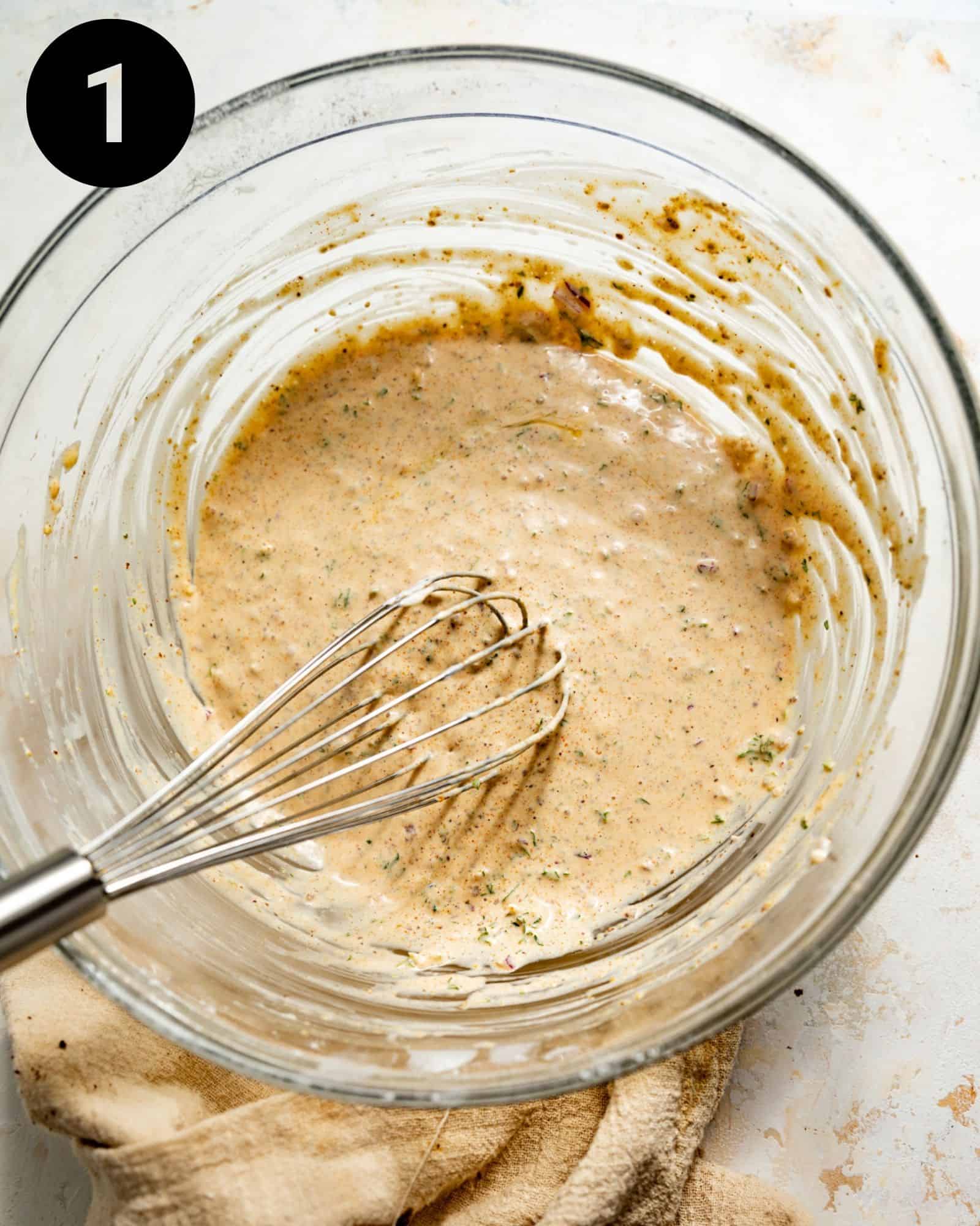 A whisk in a glass mixing bowl of mayo and seasonings to make crab cakes.