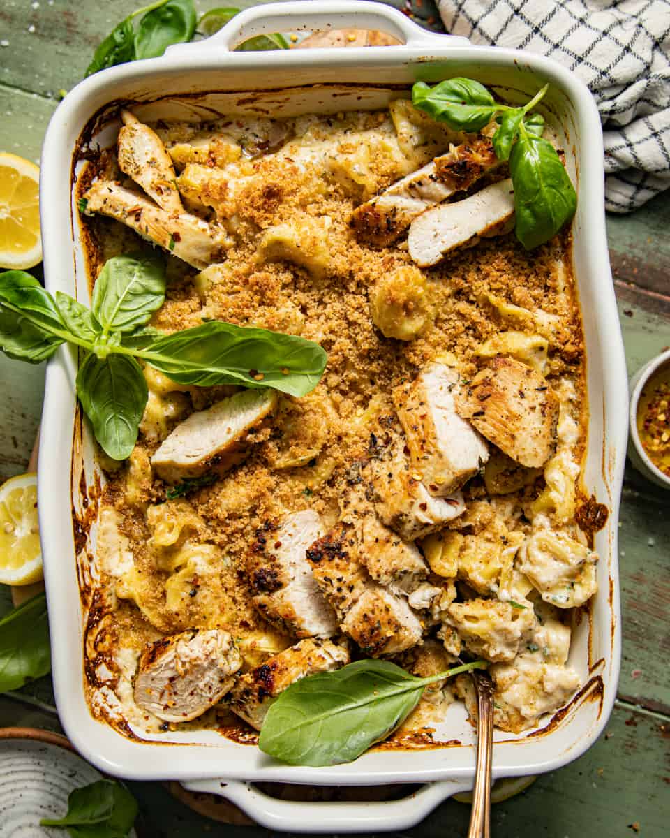 asiago tortelloni alfredo with grilled chicken in a baking dish garnished with fresh herbs and topped with sliced grilled chicken.