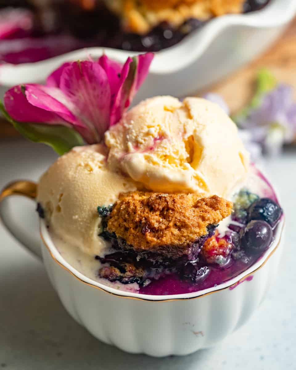blueberry cobbler in a small cup topped with ice cream and garnished with edible flowers.