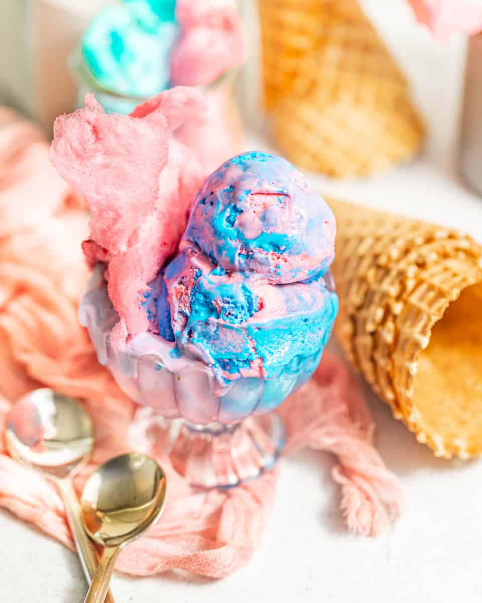 cotton candy ice cream in a glass serving cup with spoons and waffle cones behind it.