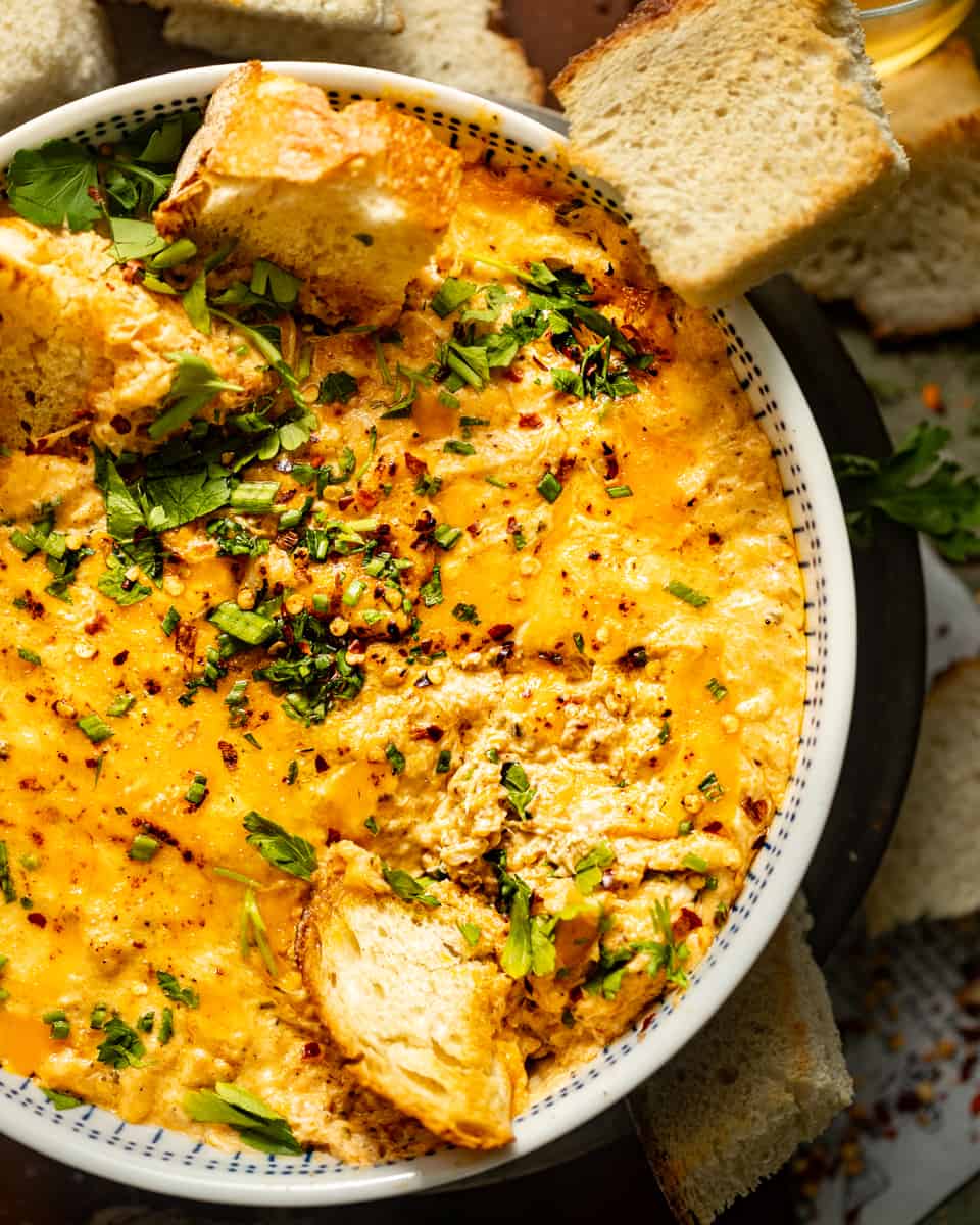 hot crab dip in a bowl with fresh herbs, bread, and red pepper flakes.