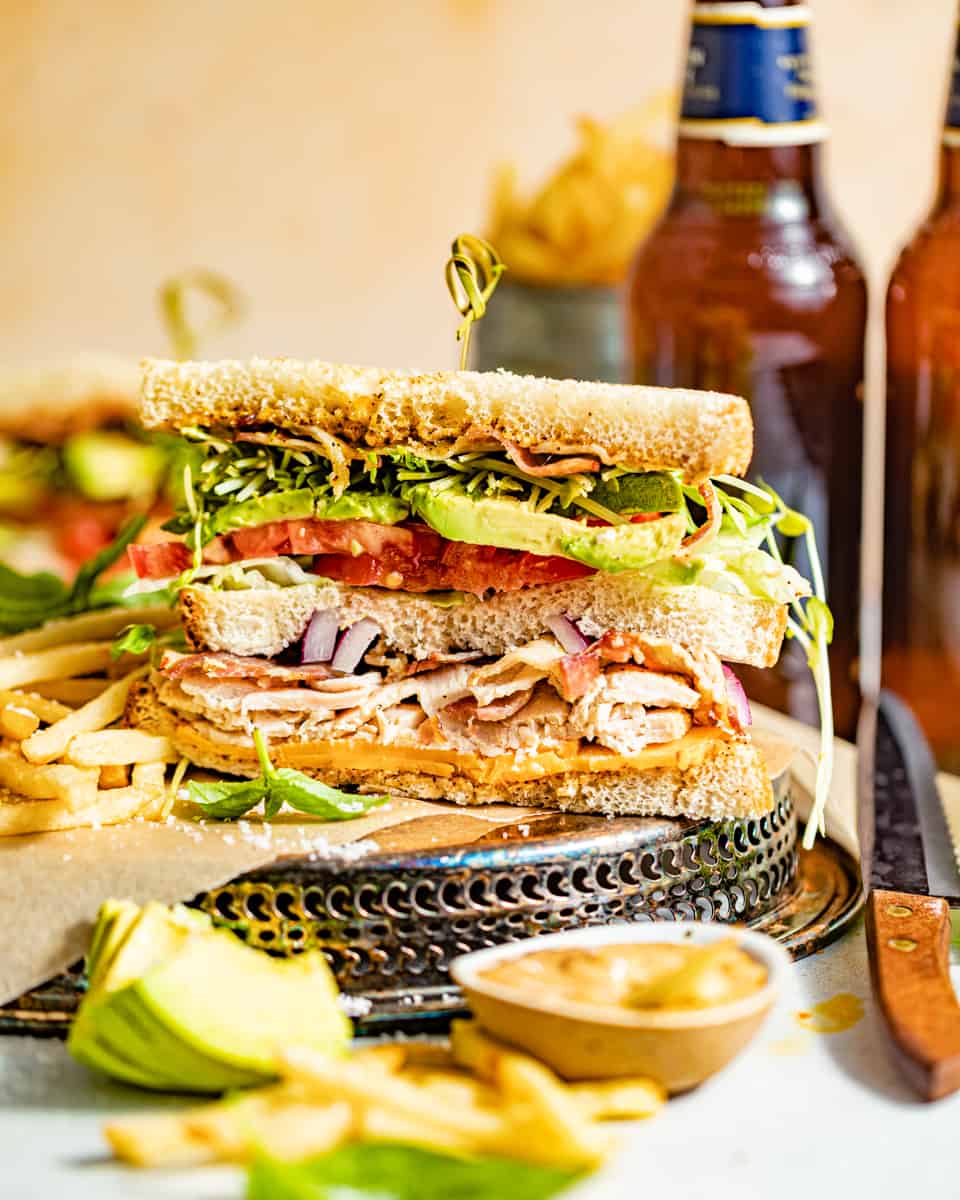 california club sandwich cut in half in front of two cans of soda and slices of avocado in front of the sandwich.