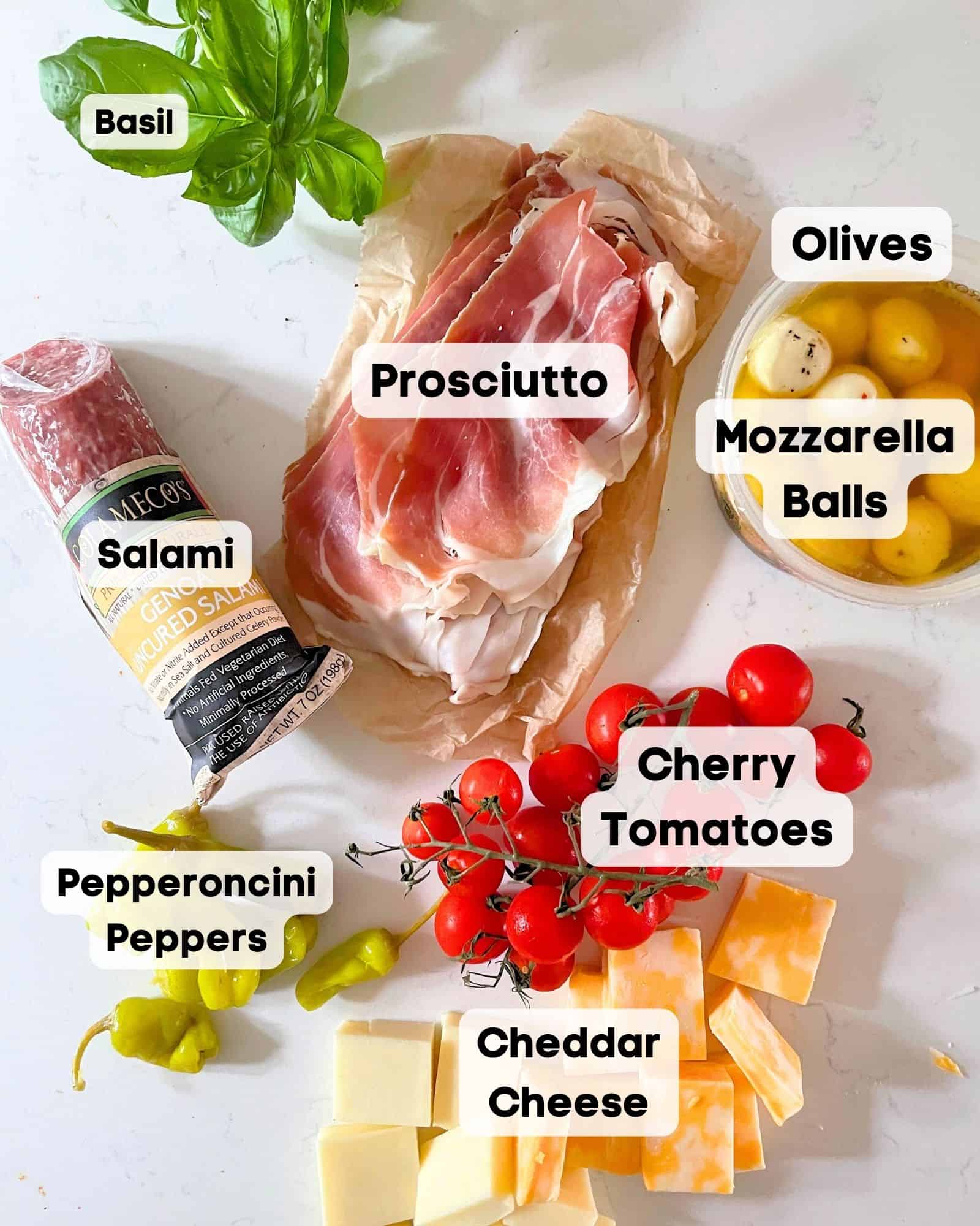 ingredients to make charcuterie skewers - prosciutto, cheddar cheese, basil, pepperoncini peppers, salami, cherry tomatoes, olives, mozzarella balls.