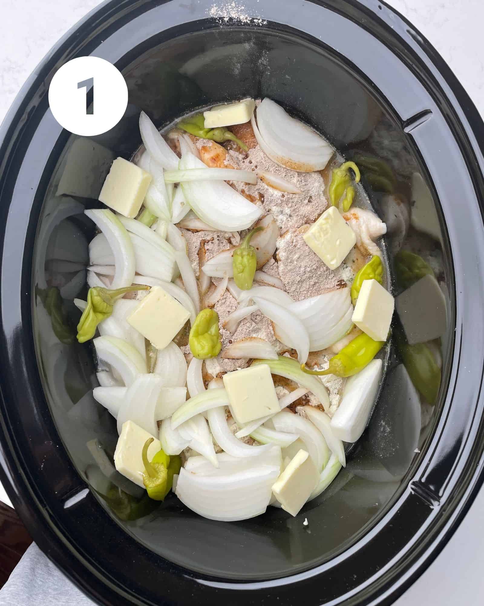 chicken in a slow cooker topped with butter, broth, ppperoncini peppers, onion, and seasoning packets.