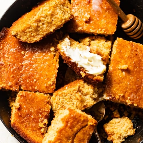 honey butter cornbread cut into pieces in a cast iron skillet with a honey dipper.
