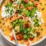 loaded baked potato soup in a bowl with a spoon and topped with crispy bacon, green onions, chives, cheddar cheese, and sour cream